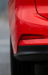 Glossy red rear bumper featuring a horizontal reflector, circular sensor, and textured matte black plastic trim.